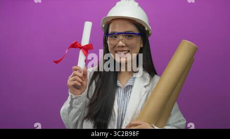 Architektin lächelt mit Blaupausen und Diplom über rosa Hintergrund, trägt Helm und Brille und zeigt beruflichen Erfolg und Erfolg Stockfoto