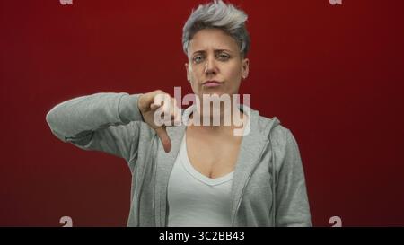 Frau mit kurzen silbernen Haaren in grauem Hoodie mit Daumen-nach-unten-Geste im roten Studio; Ablehnungsfrustration. Stockfoto