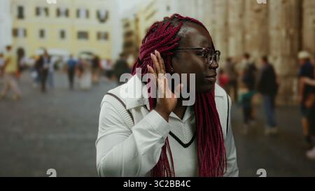 afroamerikanische Frau mit roten Zöpfen, die auf einer belebten Straße lauschen, umgeben von einem verschwommenen urbanen Hintergrund, die Outdoor-Aktivitäten und zeigt Stockfoto