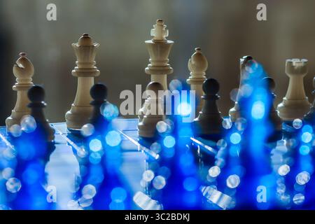 Eine dramatische Schachszene besteht aus Holzstücken auf reflektierender blauer Oberfläche und sanften Bokeh-Lichtern, die Tiefe und Intrige verleihen Stockfoto