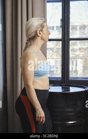 Eine Frau steht neben einem Fenster, trägt einen Sport-BH und Leggings und blickt nachdenklich nach draußen. Der Raum ist gut beleuchtet und hebt ihre fokussierten Expressi hervor Stockfoto
