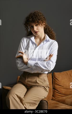 Eine junge Frau mit lockigen Haaren sitzt auf einem Sofa, trägt ein gestreiftes Hemd und eine beige Hose Stockfoto