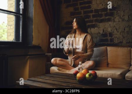 In einem gemütlichen Zuhause sitzt eine schwangere Frau bequem auf einer Couch mit einem Glas Mineralwasser und blickt aus dem Fenster Stockfoto