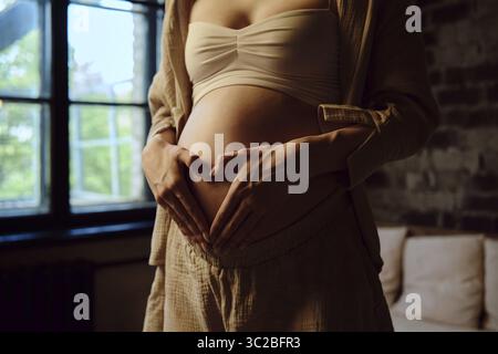 In einem gemütlichen Zuhause steht eine schwangere Frau neben einem Fenster und legt ihre Hände sanft in Herzform auf ihren abgerundeten Bauch. Sanfte Leuchte leuchtet auf Stockfoto