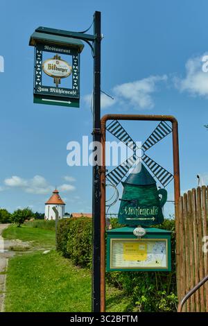 11. Juni 2025 – Woldegk-Deutschland: Grünes, schmiedeeisernes Schild in Form einer Windmühle markiert den Eingang zum Mühlencafé, mit einer Speisekoffer und weiß Stockfoto
