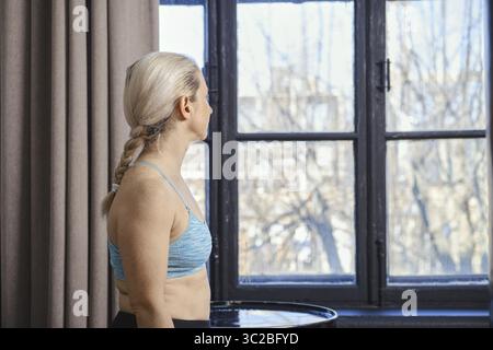 Eine Frau mit geflochtenen Haaren steht neben einem großen Fenster und blickt nach draußen auf den hellen Tag. Sie trägt ein blaues Sporttop und wirkt nachdenklich, Su Stockfoto