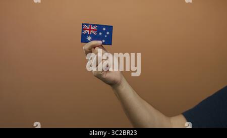 Ein Mann hält eine kleine australische Flagge an der braunen Wand mit Fokus auf Stickdetails und zeigt Nationalstolz und Identität in einem einfachen, isolierten Set Stockfoto