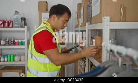 Junger Mann im Wohltätigkeitszentrum, der Spenden in einer gut sichtbaren Weste mit Tablet organisiert und sich auf protokollierte Gegenstände für den Vertrieb und die Verwaltung konzentriert Stockfoto