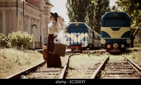 Eine Frau in Vintage-Kleidung steht auf der Eisenbahn mit Koffer in der Nähe von zwei blauen Zügen Stockfoto