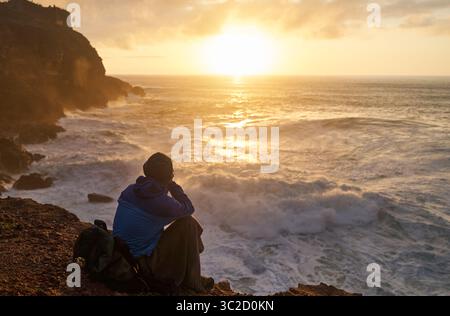 Die Person sitzt auf einer Klippe und blickt auf das Meer, während die Sonne über Wasser, Himmel und Horizont untergeht Stockfoto