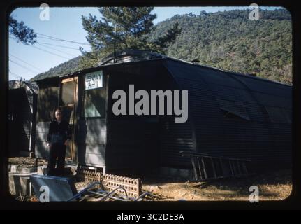 Ein Seemann der US Navy steht vor der „US NAVY A-1“, einer Hütte im Quonset-Stil, die sich im Februar 1957 auf einem bewaldeten Hügel in Chinhae, Südkorea, befindet. Das Gebäude diente während der Wiederaufbauphase nach dem Koreakrieg als Teil der Wohnräume für die U.S. Naval Advising Group. Diese Farbfolie bietet eine seltene Darstellung des amerikanischen Militärlebens während der Beratungsphase der Beziehungen zwischen den USA und Südkorea. Die bewaldete Landschaft und die militärische Infrastruktur spiegeln die Lage einer wichtigen Marinestadt wider. Stockfoto