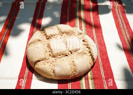 Rustikales, handgemachtes Rundbrot mit dekorativer Einkerbung, auf einem traditionellen gestreiften Tuch unter natürlichem Sonnenlicht platziert. Hausgemacht, einfach und authentisch. Stockfoto
