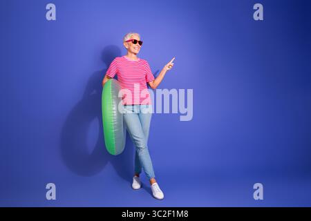 Ältere Frau in lässiger Kleidung, die selbstbewusst mit einem lustigen aufblasbaren Ring vor einem hellen violetten Hintergrund Gesten gibt Stockfoto