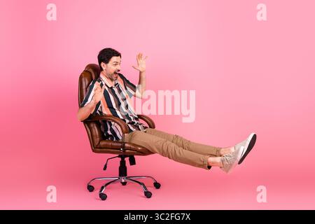 Lächelnder Mann in einem lässigen gestreiften Hemd, der einen verspielten Moment auf einem Stuhl vor einem leuchtend rosa Hintergrund genießt Stockfoto