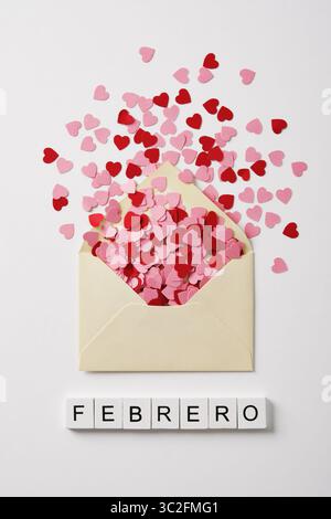 Heller Umschlag mit Konfetti-Herzen, die aus ihm herausfliegen. Das Wort Febrero steht auf weißen Holzblöcken, Februar auf Spanisch. Stockfoto