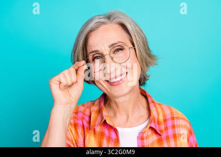 Reife Frau mit fröhlichem Ausdruck in trendiger Kleidung und Brille vor türkisfarbenem Hintergrund, die Charme ausstrahlt Stockfoto