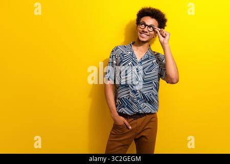 Lächelnder junger Mann mit lockigen Haaren, der ein gemustertes blaues Hemd trägt und sich in fröhlichem Casual-Stil vor gelbem Hintergrund posiert Stockfoto