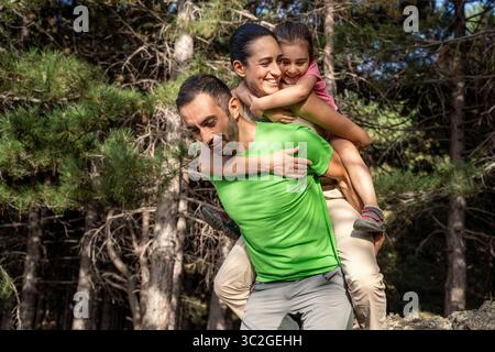 Eine glückliche und verspielte Familie genießt einen Tag im Wald, wobei ein starker Vater sowohl seiner lächelnden Frau als auch ihrer fröhlichen Tochter einen Huckepack-Ritt gibt Stockfoto