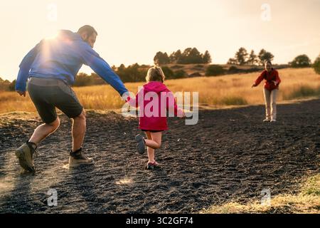 Adult and child holding hands while running toward a woman - Family outdoor adventure - joyful reunion in nature at sunset. Stockfoto