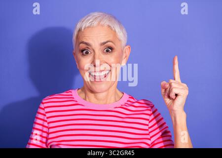 Lächelnde Frau, die mit ihrem Finger einen verspielten Gedanken vor einem leuchtend violetten Hintergrund in stilvoller Kleidung macht Stockfoto