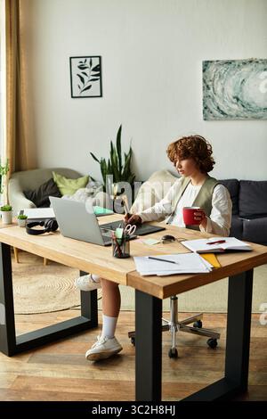 Eine junge Frau mit lockigen Haaren konzentriert sich auf ihre Arbeit, während sie einen Kaffee in ihrer gemütlichen Wohnung genießt. Stockfoto