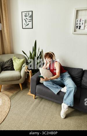 Eine Frau mit lockigen Haaren sitzt auf einer Couch, taucht in ihr Handy ein, während sie sich in ihrer Wohnung auszieht. Stockfoto