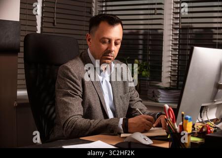 Selbstbewusster Geschäftsmann in einem klassischen grauen Blazer, der sich auf die Arbeit an einem hellen Schreibtisch konzentriert. Moderner Arbeitsbereich, Jalousien an den Fenstern, Drucker und Monitore. Stockfoto
