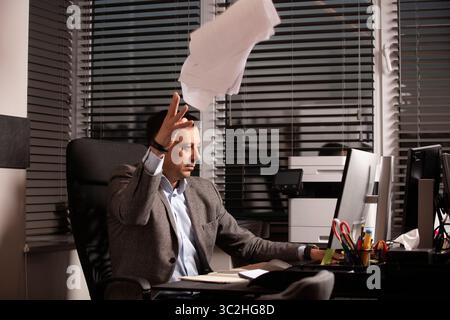 Ein gestresster Büroangestellter tippt auf einem Laptop, während die Papiere um ihn herum fliegen. Überforderte Geschäftsatmosphäre, dringende Termine. Multitasking und Paperchaos. Stockfoto