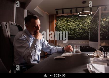 Lächelnder Geschäftsmann mittleren Alters, der sich an seinem Schreibtisch in einem modernen, umweltfreundlichen Büro entspannt. Warmes Licht, stilvolles Interieur und lässige Stimmung. Stockfoto