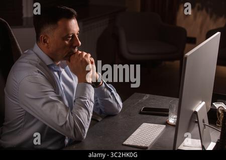 Ein fokussierter Mann in einem leichten Hemd hält das Smartphone, während er in einem stilvollen Abendbüro arbeitet. Moderner Arbeitsbereich mit grüner Wand und eleganten Beleuchtungssets. Stockfoto