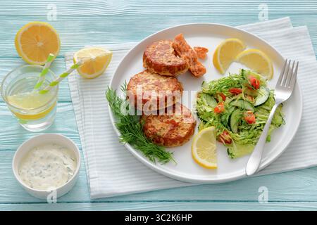 Geräucherte Lachsburger, Pasteten oder Kuchen, serviert mit Krautsalat und Zitronenscheiben auf einem weißen Teller auf Holztisch mit Tzatziki-Sauce und Sprudelwasser, Stockfoto