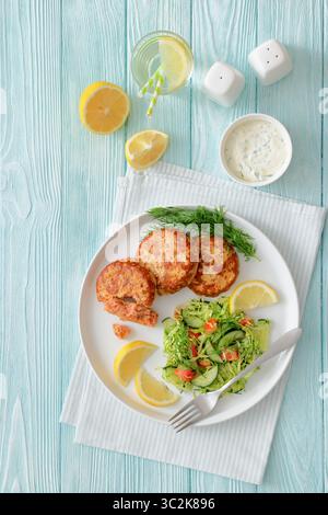 Geräucherte Lachsburger, Pasteten oder Kuchen serviert mit Krautsalat und Zitronenscheiben auf einem weißen Teller auf Holztisch mit Tzatziki-Sauce und Wasser, vertikal Stockfoto