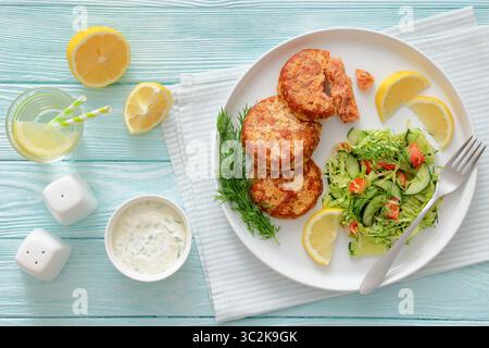 Geräucherte Lachsburger, Pasteten oder Kuchen serviert mit Krautsalat und Zitronenscheiben auf einem weißen Teller auf cyanfarbenem Holztisch mit Tzatziki-Sauce und Wasser Stockfoto