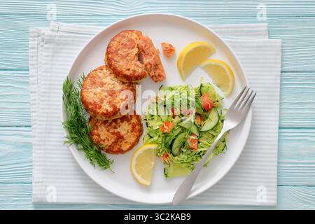 Geräucherte Lachsburger, Pasteten oder Kuchen serviert mit Krautsalat und Zitronenscheiben mit Gabel auf weißem Teller auf cyanfarbenem Holztisch, Blick von oben, Stockfoto