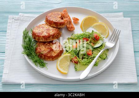 Geräucherte Lachsburger, Pasteten oder Kuchen serviert mit Krautsalat und Zitronenscheiben mit Gabel auf weißem Teller auf cyanfarbenem Holztisch, Nahaufnahme, nicht AI Stockfoto