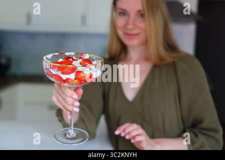 Glückliche junge Frau genießt hausgemachtes Erdbeerparfait in moderner Küche. Stockfoto