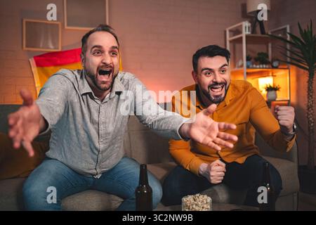 Die Spannung erfüllt den Raum, während Freunde ihr Team während eines intensiven Sportspiels zu Hause an einem gemütlichen Abend anfeuern Stockfoto