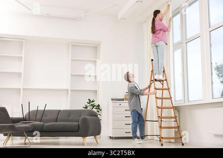 Ein Paar, das Vorhänge mit einer Leiter im Zimmer aufstellt Stockfoto