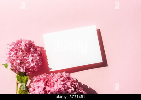 Leere weiße Karte mit rosa Hortensie-Blüten auf pastellrosa Hintergrund mit harten Schatten. Draufsicht, flach, Modell. Stockfoto