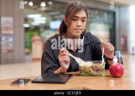 Reife Transgender Frau essen gesunden Salat mit Apfel und Wasser in zwangloser Mall Restaurant Umgebung Stockfoto