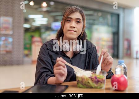 Reife Transgender Frau essen gesunden Salat mit Apfel und Wasser in zwangloser Mall Restaurant Umgebung Stockfoto