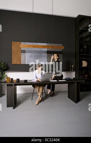 Zwei Frauen nehmen an einem produktiven Meeting in einer eleganten Büroumgebung Teil. Einer sitzt an einem Schreibtisch und arbeitet an einem Laptop, während der andere in der Nähe eines steht Stockfoto