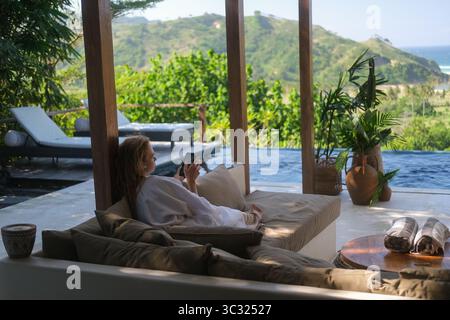 Eine junge Frau im Bademantel sitzt auf der Terrasse einer Luxusvilla mit Pool Stockfoto