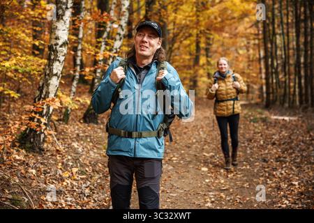 Zwei Rucksacktouristen spazieren auf dem Wanderweg im Herbstwald. Wanderfreunde in der Herbstsaison im Wald. Der lächelnde Mann trägt eine wasserdichte Jacke Stockfoto