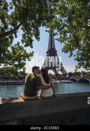 Ein Paar, das auf der Wand sitzt, küsst sich mit dem Eiffelturm in der Ferne in Paris. Stockfoto