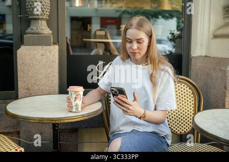 Junge Frau trinkt Kaffee im Café im Freien und benutzt Smartphone Stockfoto