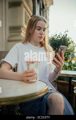 Junge Frau trinkt Kaffee im Café im Freien und benutzt Smartphone Stockfoto
