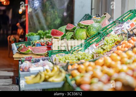 Verschiedene frische Früchte wie Wassermelonen, Trauben, Bananen, Orangen und Beeren werden abends an einem Marktstand in einer urbanen Umgebung im Freien serviert. Stockfoto