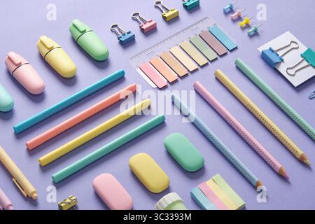Schreibwarenhintergrund mit pastellfarbenen Schreibwaren, die ordentlich auf einem hellvioletten Hintergrund angeordnet sind. Stockfoto