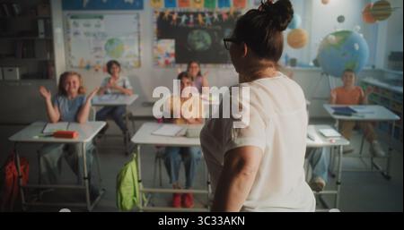 Fröhliche, abwechslungsreiche Kinder sitzen an Schreibtischen, klatschen die Hände, interagieren mit dem Lehrer während der dynamischen Gamified-Aktivität. Lehrerin, die Grundschüler interaktiven Unterricht gibt. Stockfoto
